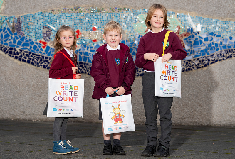 Three pupils in uniform holding their Read, Write, Count and Bookbug bags